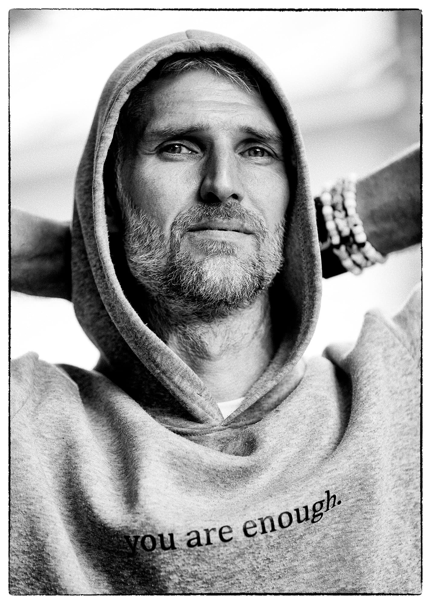 Dominic dans son coton ouaté gris, capuchon relevé, les mains derrière la tête, le regard vers le haut. Sur la poitrine, en petits caractères : you are enough. Son propre tirage en noir et blanc.