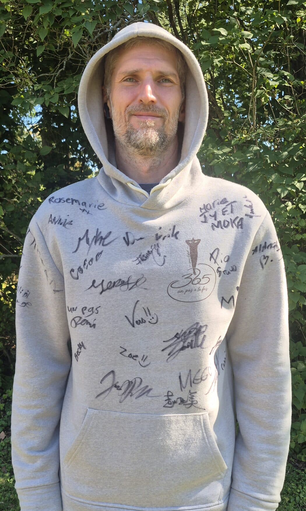 Dominic in the signed hoodie, hood up, half smiling, standing in front of the trees. The front is covered in signatures around the small 365 logo on the chest.